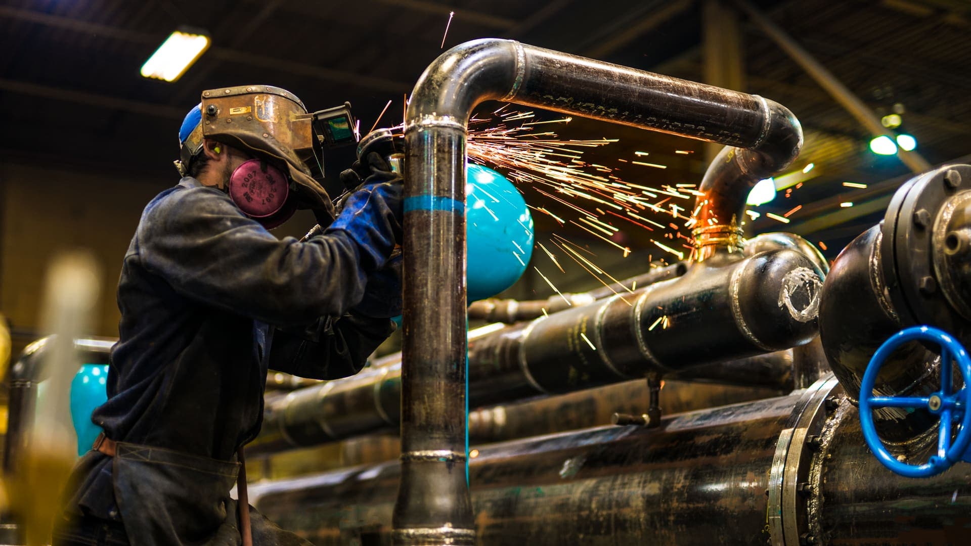 Tradesperson in protective gear grinding a steel pipe, sparks flying